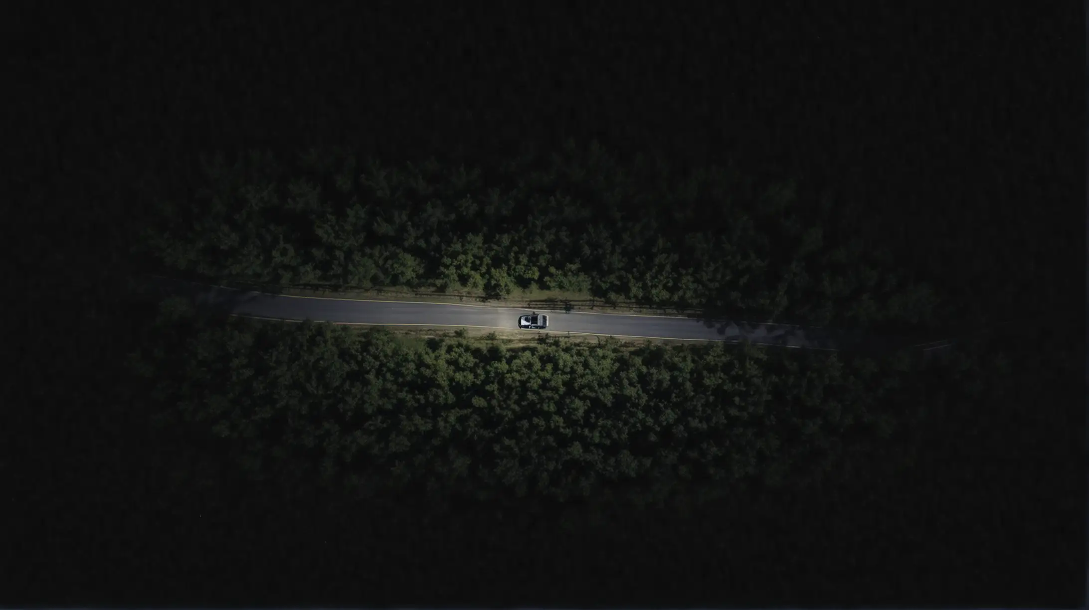 Generate a very high-angle overhead shot of a vehicle traveling down a dark, winding, rural southern road at night.